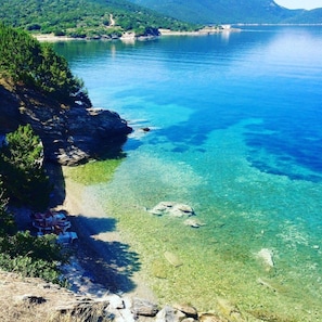 A clear coastal view with a rocky shoreline, a small beach area with lounge chairs, and a distant mountain range. - Venus Beach Hotel (Karystos)