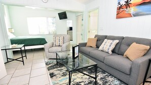 A living room with a grey sofa, patterned armchair, and a glass coffee table. A bed with green bedding is visible in the background. - Morningside (Sanibel)