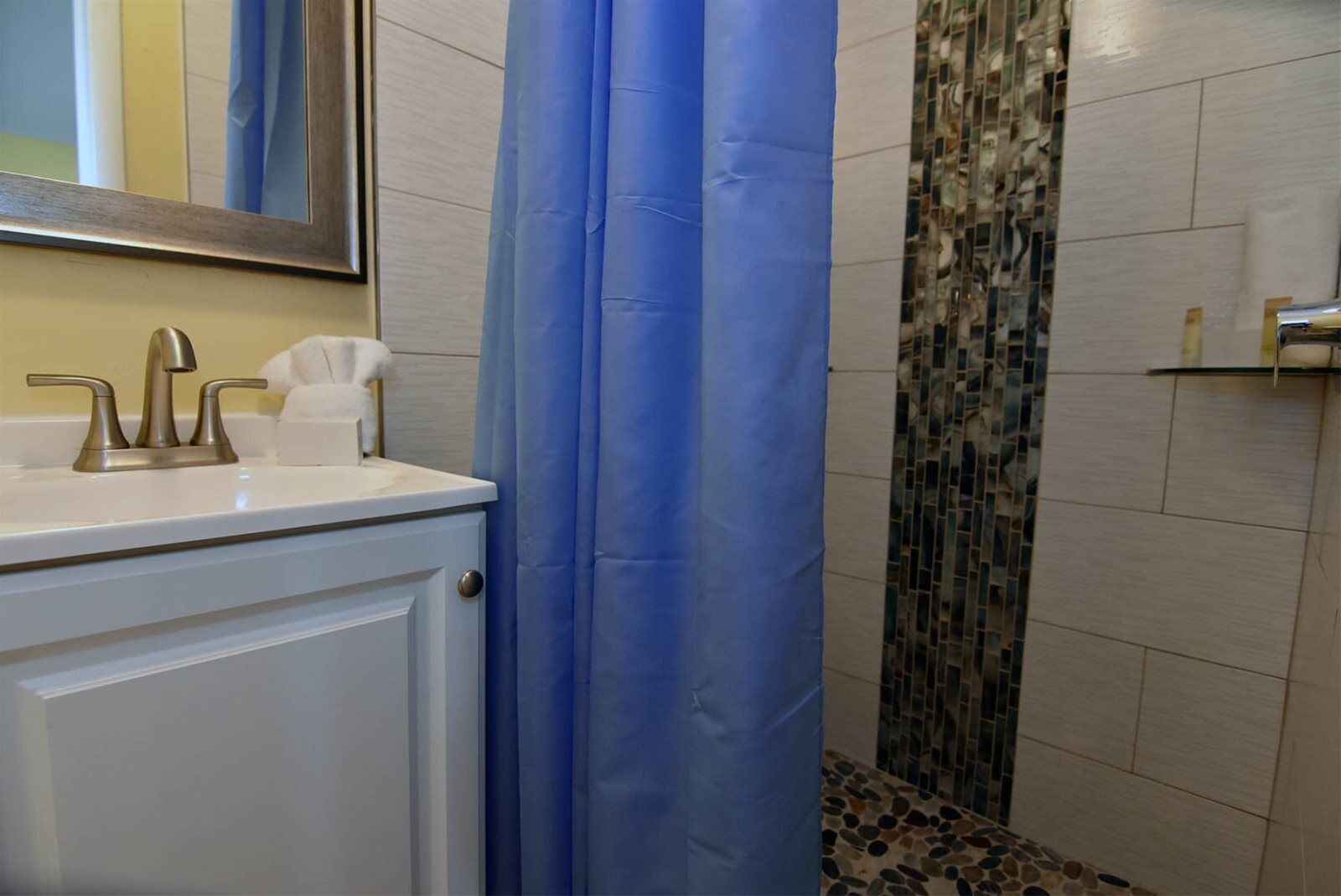 A bathroom with a white vanity, a mirror, a blue shower curtain, and a mosaic tile shower wall.