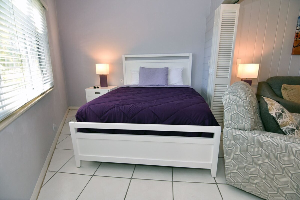A bedroom with a white bed, purple bedding, a nightstand with a lamp, and a patterned armchair.
