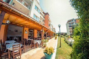 Un área de café al aire libre con mesas y sillas, rodeada por un paseo techado y un jardín con un parque infantil.