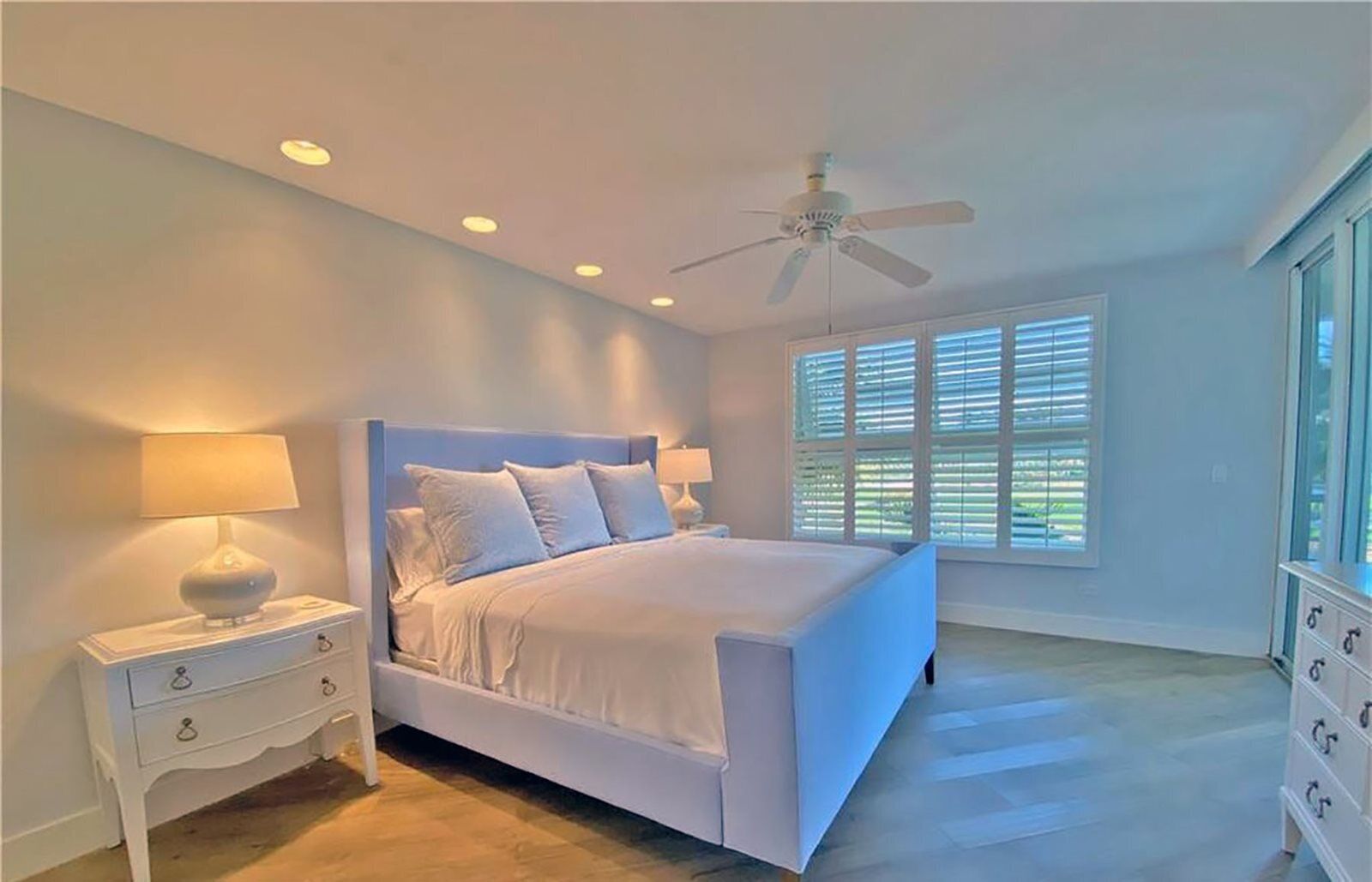 A bedroom with a large bed, two bedside tables, a ceiling fan, and a window with blinds.