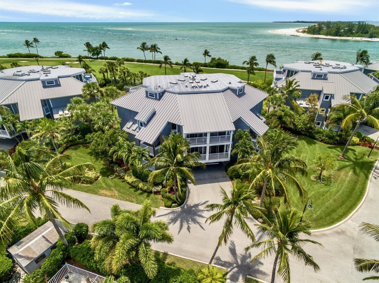 A resort with multiple buildings featuring flat roofs, surrounded by palm trees and a beach view.