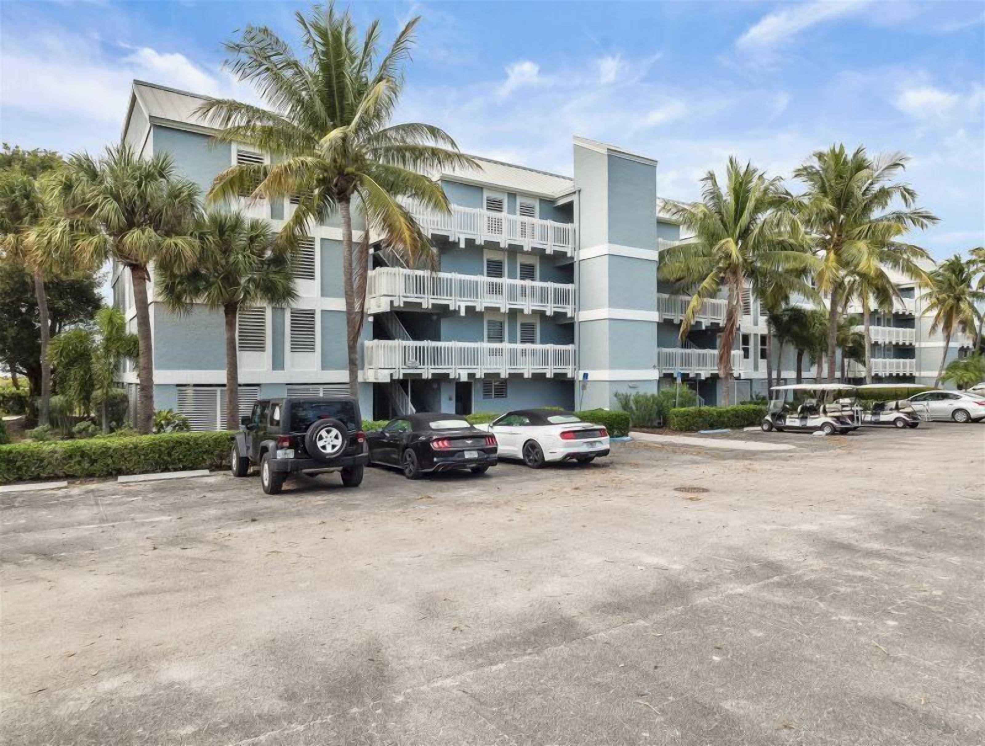 A multi-story hotel with balconies, palm trees, and a parking area with cars and golf carts.