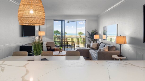A modern living room with a large window, a dining area, and a balcony with a view of the beach. - Beach Villas (Captiva)