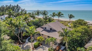 Una casa de dos pisos con techo plano, rodeada de palmeras y vista al mar.