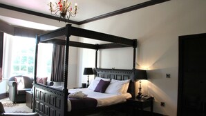 A four-poster bed with a canopy, a bedside table with a lamp, and a chair in a room with a chandelier. - The Carlton Manor Hotel (Lowestoft)