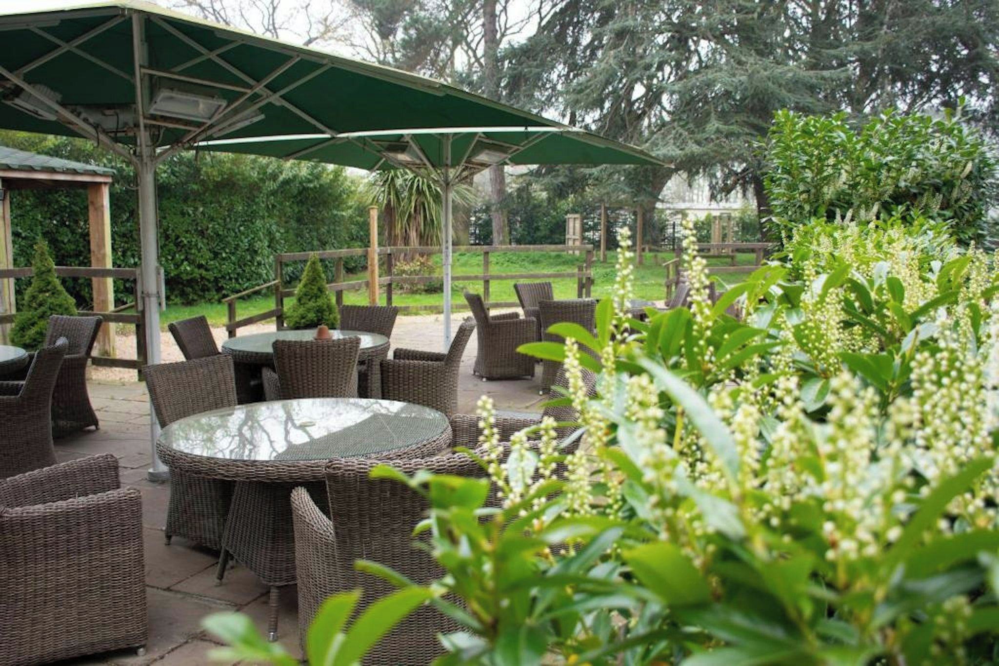 An outdoor patio with wicker furniture and a glass-top table, surrounded by greenery.
