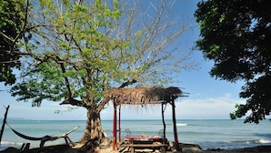 Escena de playa con una estructura de techo de paja, una hamaca y una mesa de madera con sillas.