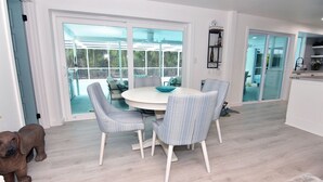 Een eetruimte met een ronde tafel en vier stoelen, grote glazen deuren die uitkomen op een uitzicht, en een zichtbare keuken achter de glazen deuren.