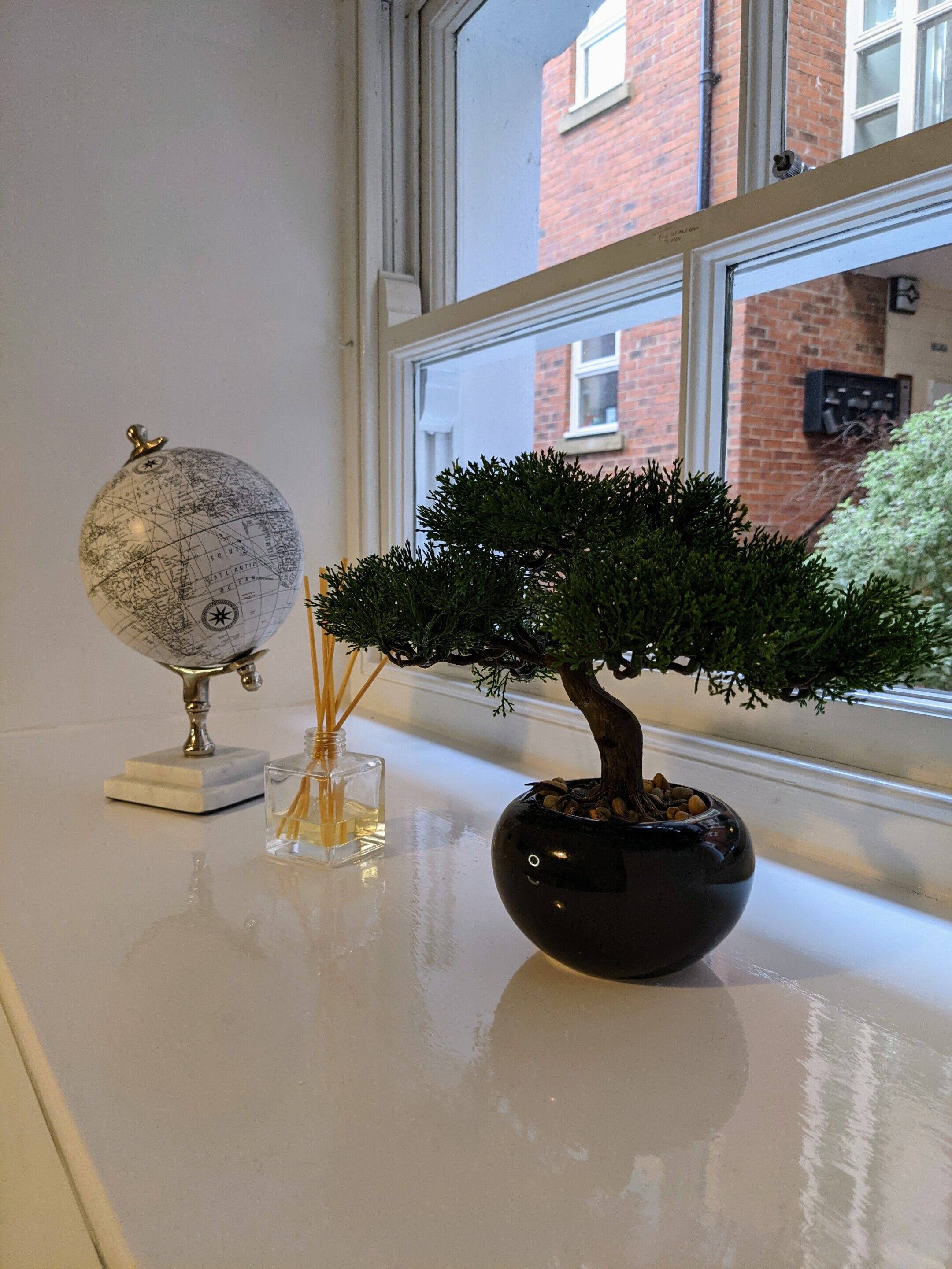 A globe on a stand, a bonsai tree in a pot, and a window with a view of a brick building.