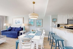 Una cuina moderna amb armaris blancs, una illa central amb tamborets i una zona de menjador amb estovell blau i blanc.