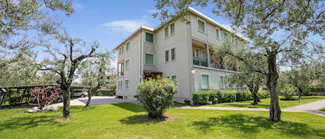 Un bâtiment à deux étages doté d’un balcon au deuxième étage, entouré d’une pelouse bien entretenue et d’arbres.