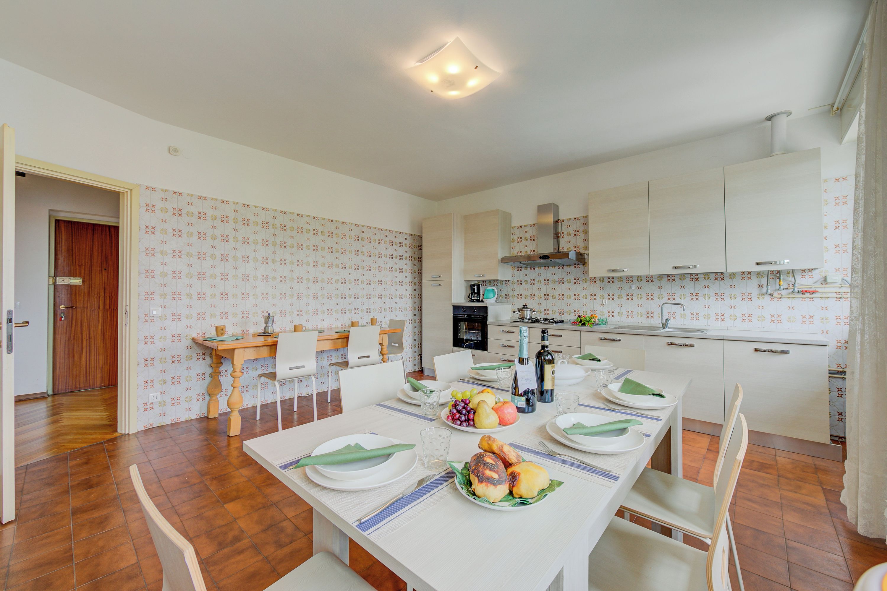 A dining area with a long table set for four, a kitchen with white cabinets, and a patterned wallpaper design.