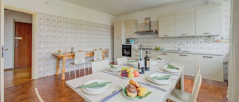 Un coin repas avec une longue table dressée pour quatre personnes, une cuisine avec des meubles blancs et un papier peint à motifs.