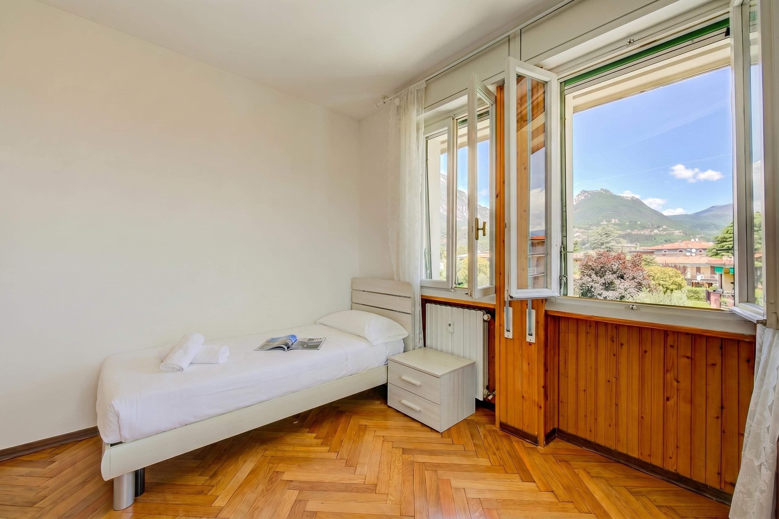A bedroom with a bed, a small bedside table, a radiator, and a window with a view of mountains.