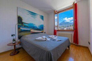 A hotel room with a bed, a painting, a window with a mountain view, and a table with a bottle and glasses.