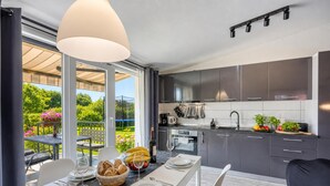 A modern kitchen with dark cabinets, a dining area with a table set for two, and a view of a garden. - Family Friendly House Kacana - Garden & BBQ (Vodnjan)