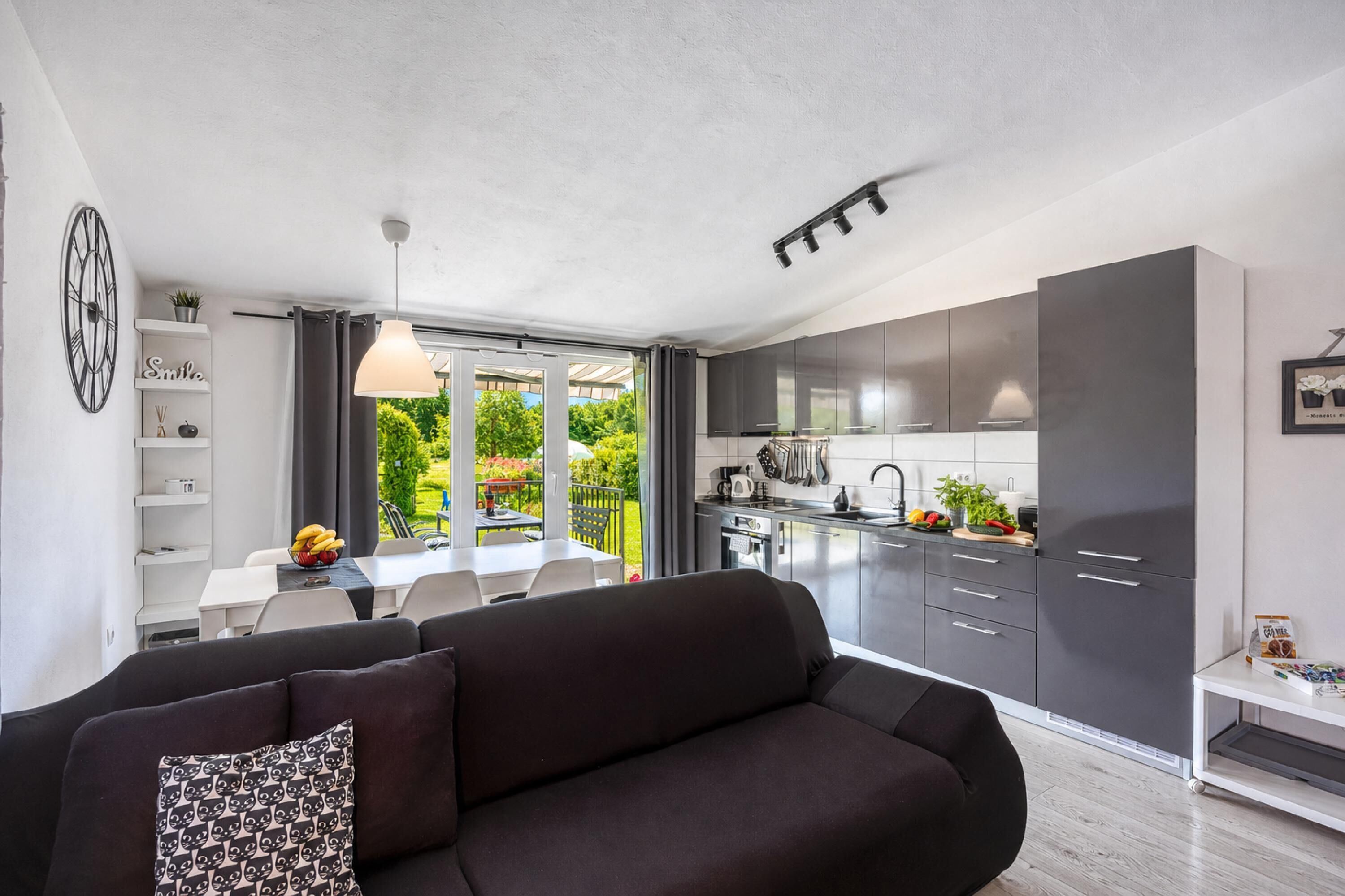 A modern kitchen with dark cabinets, a dining area with a table and chairs, and a living room with a dark sofa and patterned pillow.