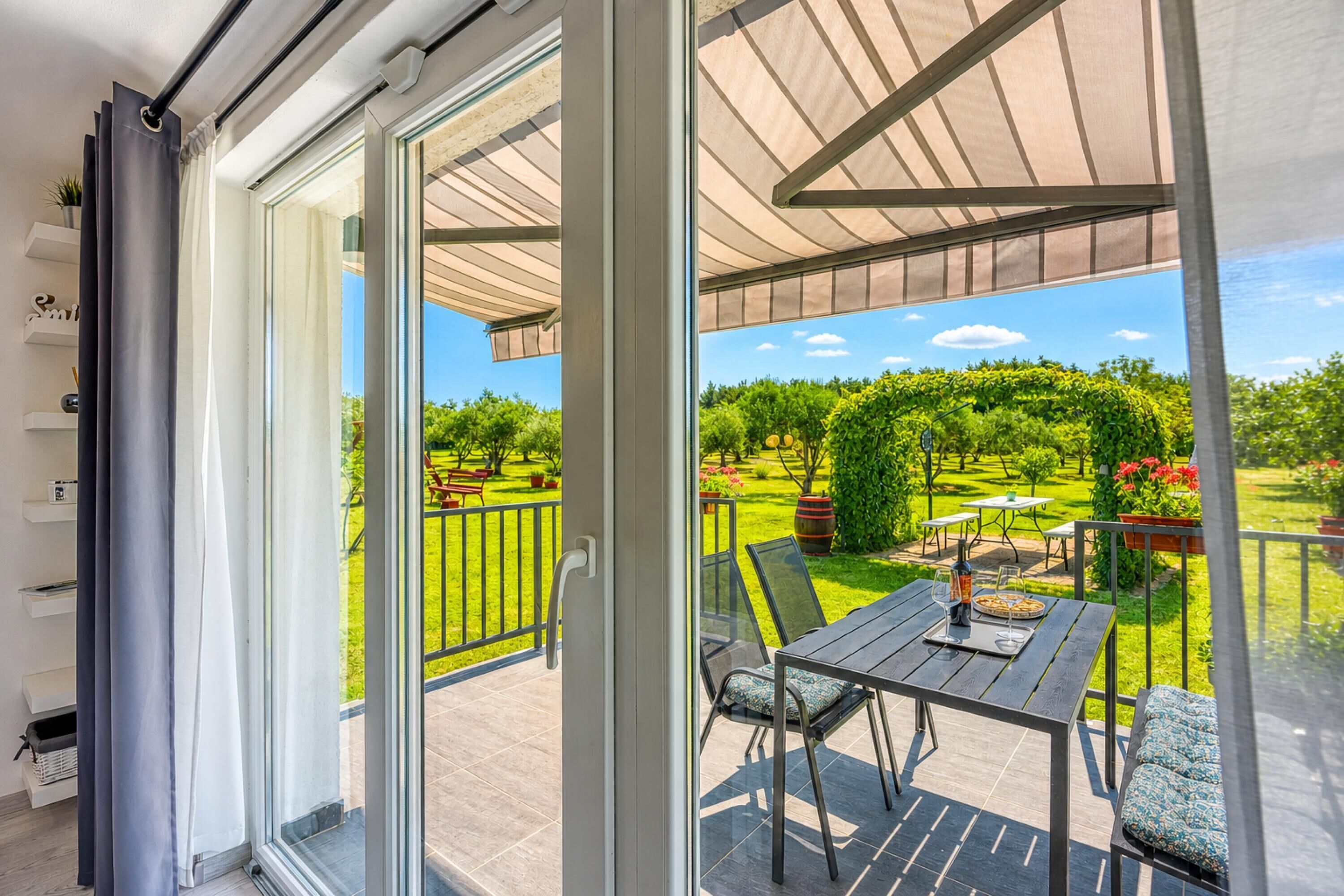A balcony with a glass door, a table with chairs, and a view of a garden.