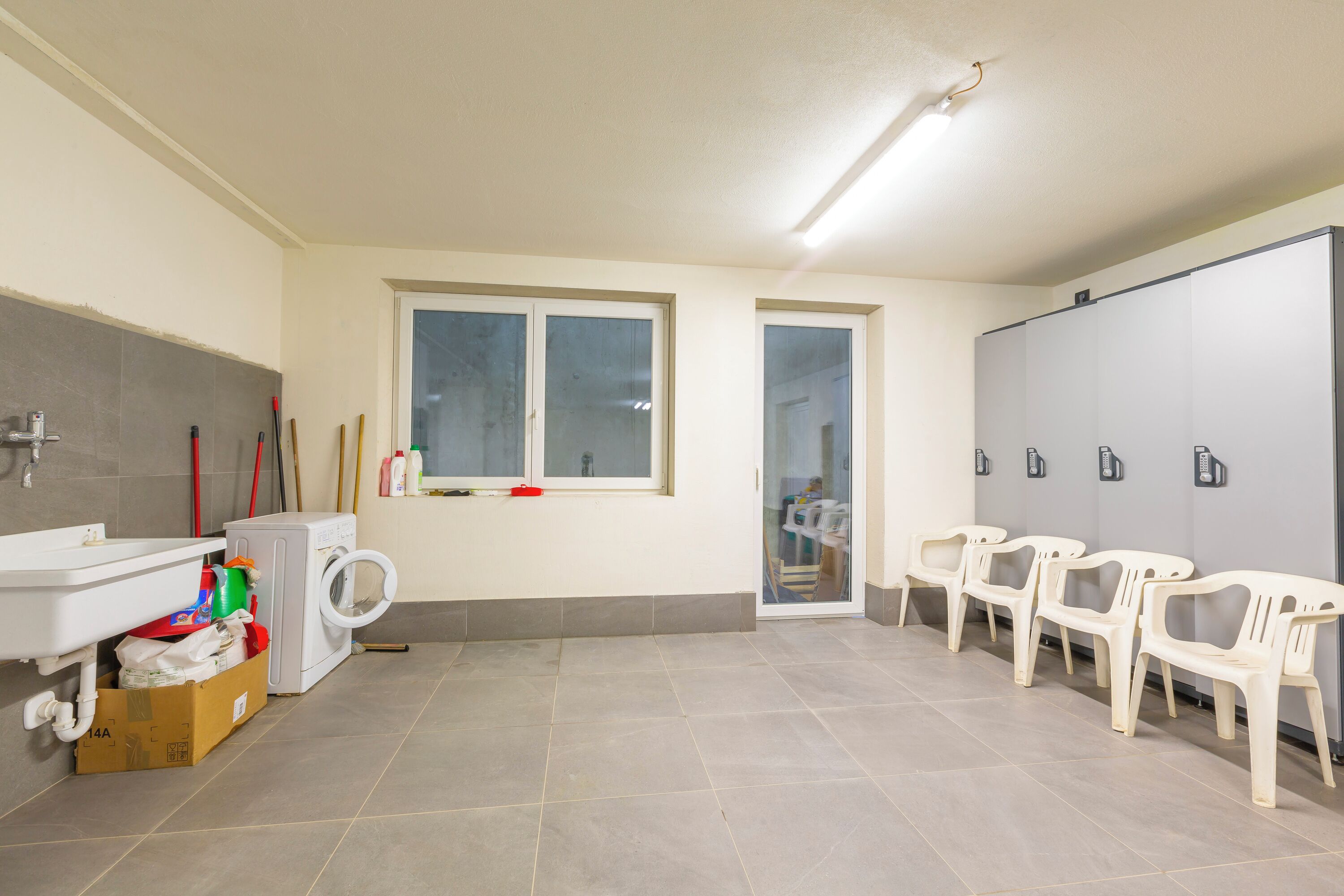 A bathroom with a sink, mirror, toilet, and a window. There are plastic chairs and a cabinet with a mirror.