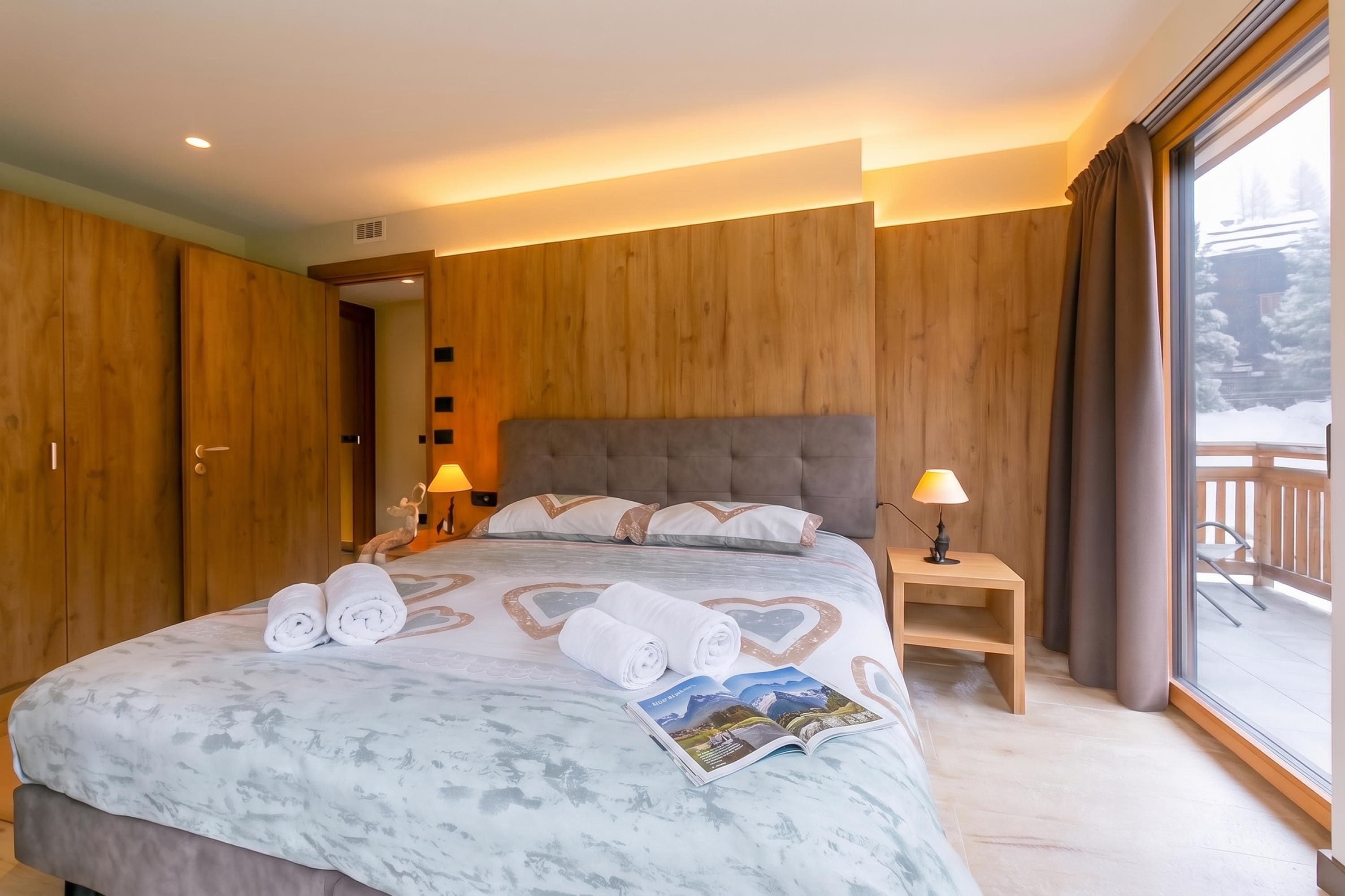 A hotel room with a large bed, wooden panel walls, two bedside lamps, and a view of a snowy landscape through a large window.