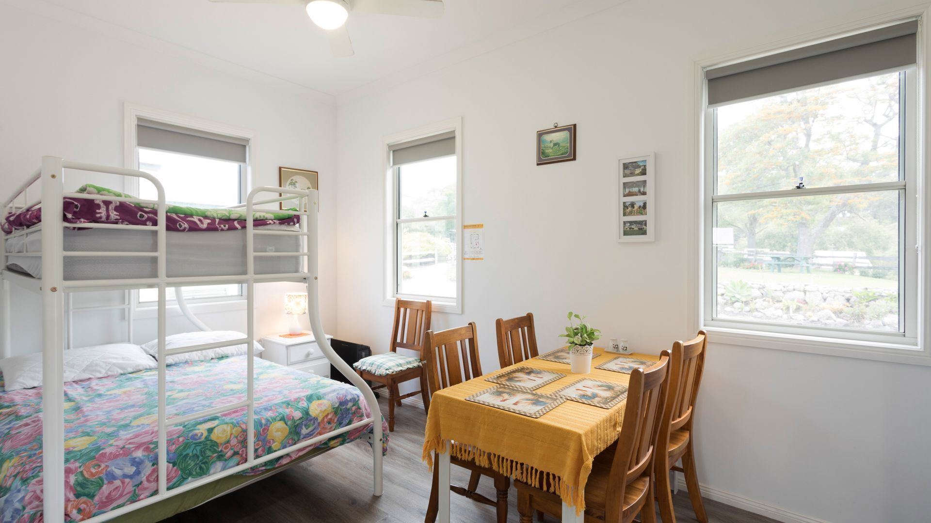 A dormitory room with bunk beds, a dining table with chairs, and a window with a view of trees.