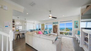 Un salon spacieux avec un grand canapé blanc, un ventilateur de plafond et une vue sur la plage grâce à de grandes fenêtres.
