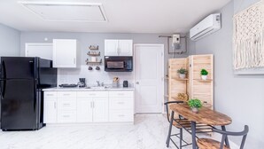 A modern kitchen with white cabinets, a black refrigerator, a microwave, and a coffee maker. There is a wooden dining table with chairs and a decorative macrame wall hanging.