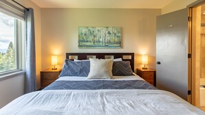 A neatly made bed with a patterned bedspread and pillows, two bedside lamps, a painting on the wall, and a door to the right. - Outpost: White Ridge A6 (Teton Village)