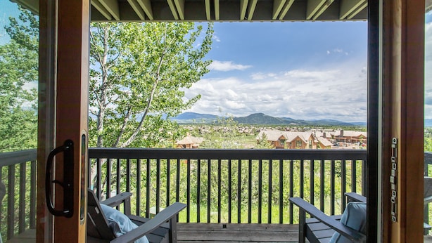 A wooden deck with two lounge chairs and a table, overlooking a scenic landscape with trees and distant buildings. - Outpost: White Ridge A6 (Teton Village)