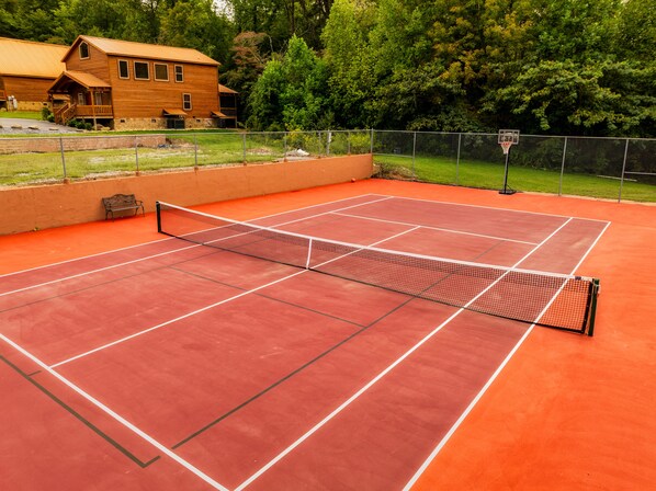 A tennis court with a net, surrounded by a fence and a wooden building in the background.