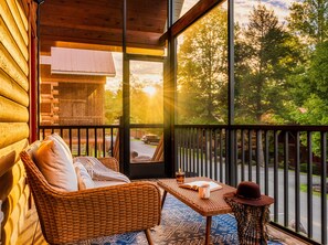 A wicker chair with a cushion and a small table with a hat and book on a balcony with a view of trees and a road.