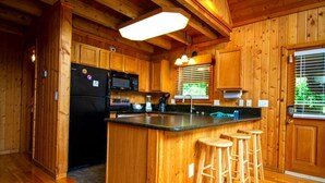 A wooden cabin-style kitchen with a central island, bar stools, a refrigerator, and a window with blinds. - Create Memories this Spring Break | Smoky Waters (Sevierville)