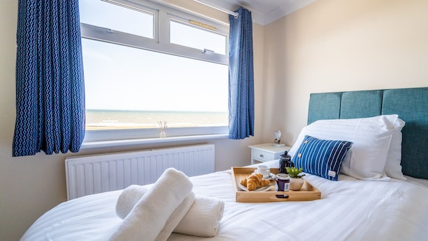 A neatly made bed with white linens, a tray with croissants and a drink, and a view of the beach through a window with blue curtains. - Sea View Penthouse (Great Yarmouth)