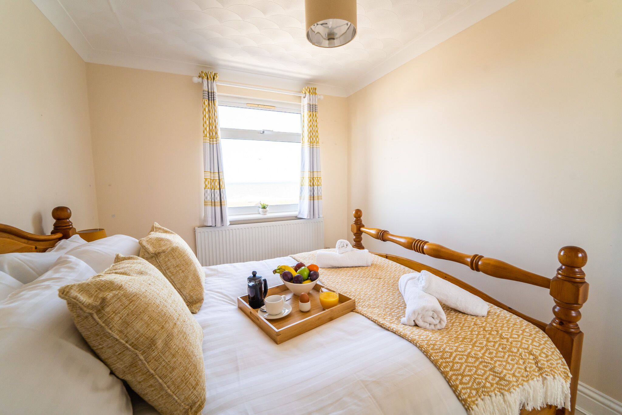 A neatly made bed with a tray of breakfast items, a window with curtains, and a radiator.