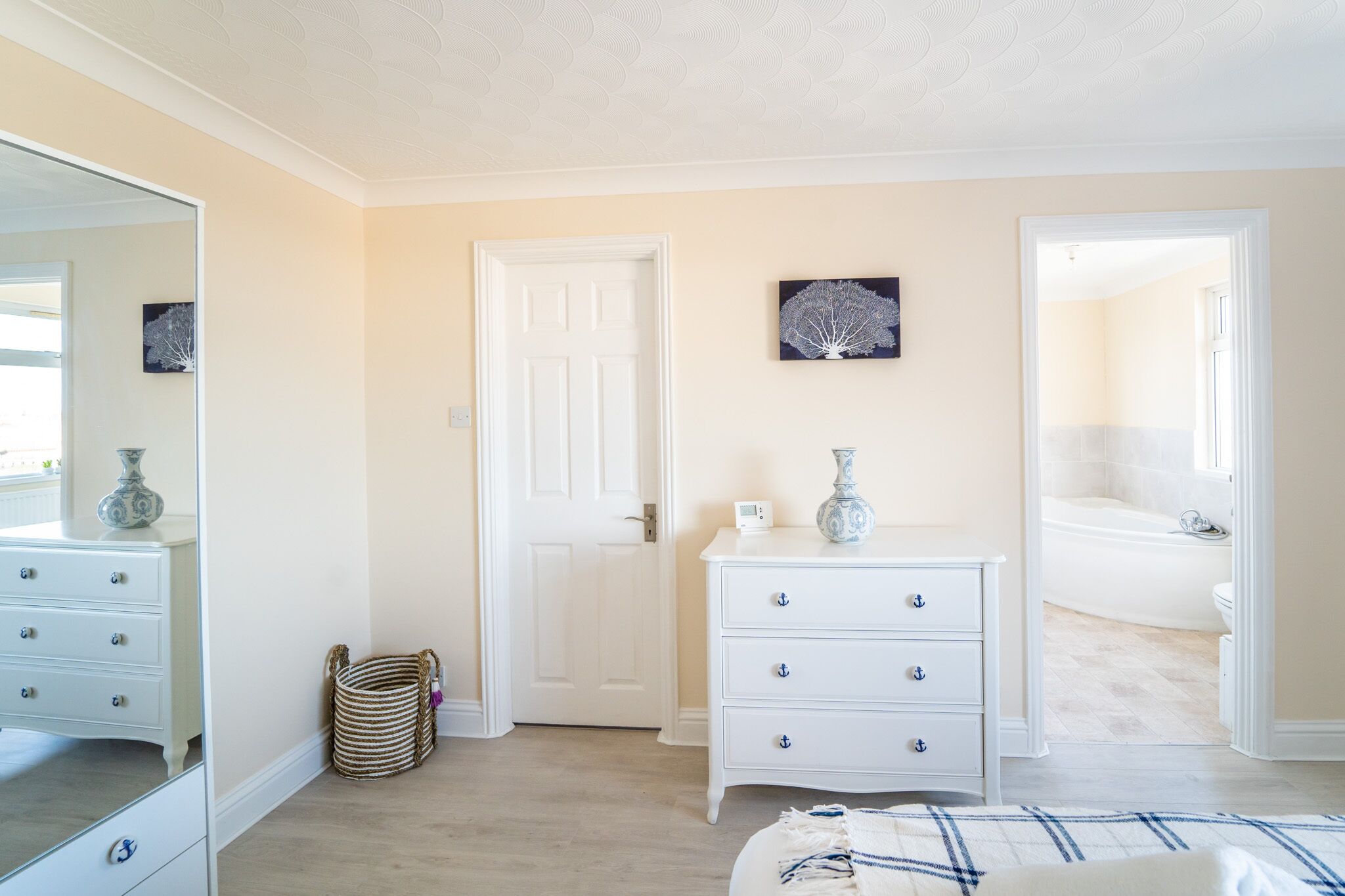 A bedroom with a large mirror, a white dresser, a bedside table, and a door leading to a bathroom.