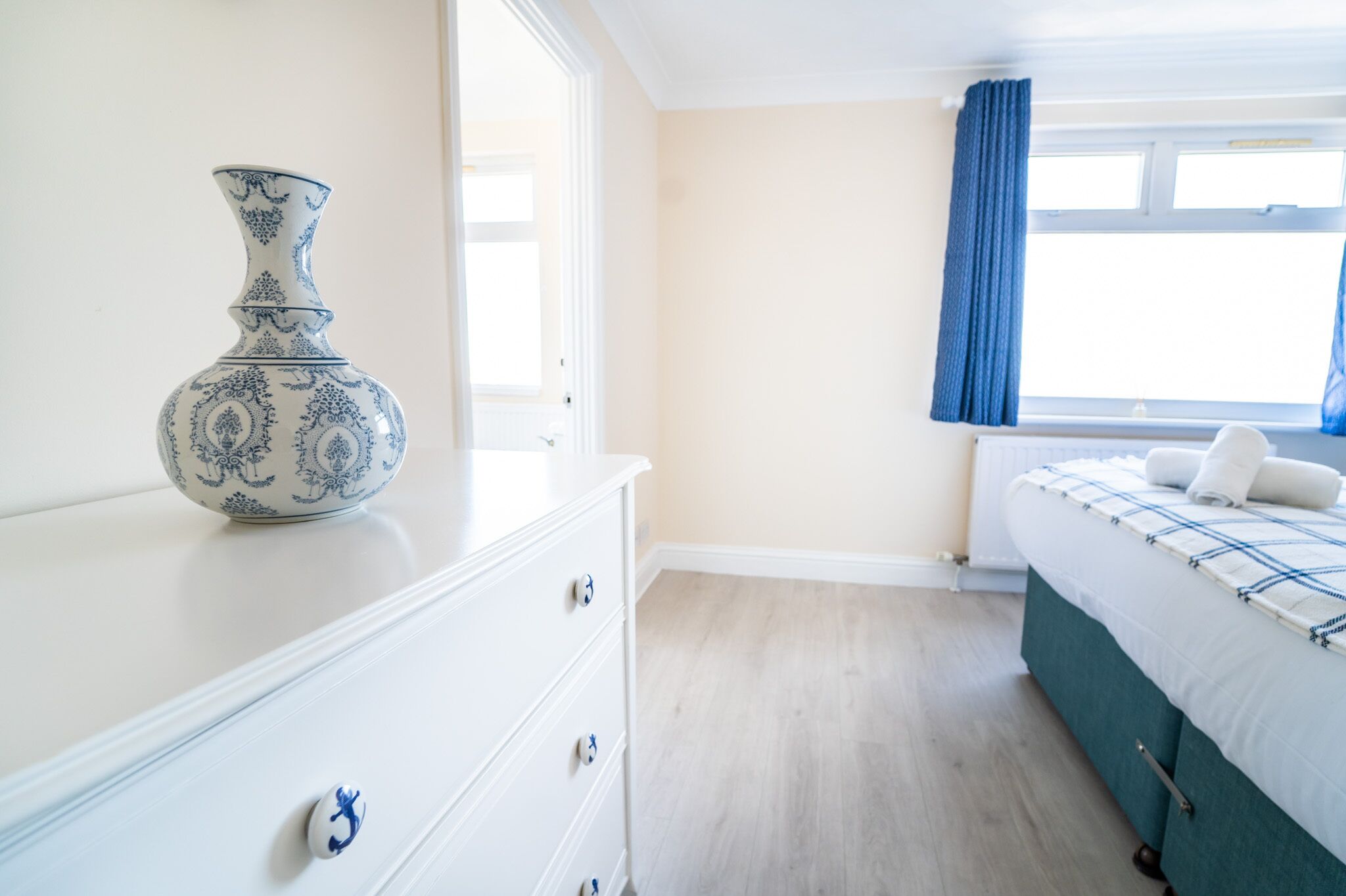 A bedroom with a white dresser, a blue and white vase, a bed with a blue and white bedspread, and a window with blue curtains.
