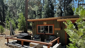 Una cabaña con base de piedra, una terraza de madera, una parrilla de barbacoa y un ventanal con un letrero que dice "The Little Cabin".