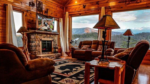 A cozy cabin interior with a stone fireplace, leather furniture, and a large window offering a mountain view. - Rustic Mountain Retreat ~ Panoramic Mountain View ~ Hot Tub ~ Game Room  (Sevierville)