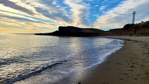 Eine Küstenansicht mit einem Leuchtturm, einem Sandstrand und felsigen Klippen unter einem bewölkten Himmel.