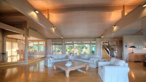 A spacious living room with a wooden coffee table, white sofas, and large windows offering a view of the outdoors. - LYCIPONON VILLA for an unforgettable, relaxing, private, and secure vacation (Ανάβυσσος)