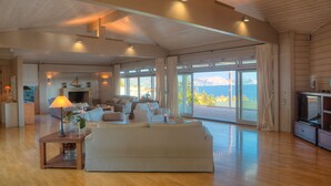 A spacious living room with a large sofa, a coffee table, and a television. There is a fireplace and a view of the outdoors through large windows. - LYCIPONON VILLA for an unforgettable, relaxing, private, and secure vacation (Ανάβυσσος)