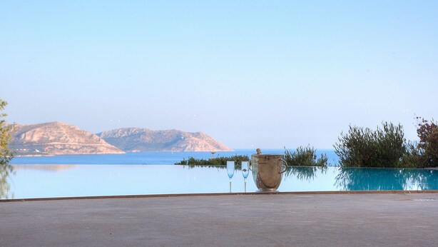 A clear view of a calm body of water with mountains in the background, a glass table with a large vase and two wine glasses, and a clear sky. - LYCIPONON VILLA for an unforgettable, relaxing, private, and secure vacation (Ανάβυσσος)