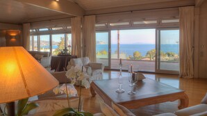 A spacious living room with a large window offering a view of the sea and hills. There is a wooden coffee table, a lamp, and white furniture. - LYCIPONON VILLA for an unforgettable, relaxing, private, and secure vacation (Ανάβυσσος)