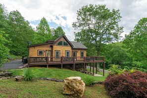 A wooden cabin with a deck, surrounded by lush greenery and trees. - Mtn Lodge~Hot Tub+Fire Pit+Fireplace, WiFi! (Burnsville)