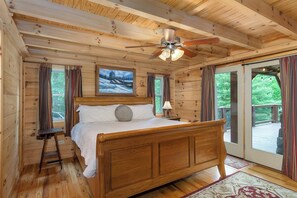 A wooden cabin bedroom with a bed, a ceiling fan, a painting, and a view through the glass door. - Mtn Lodge~Hot Tub+Fire Pit+Fireplace, WiFi! (Burnsville)