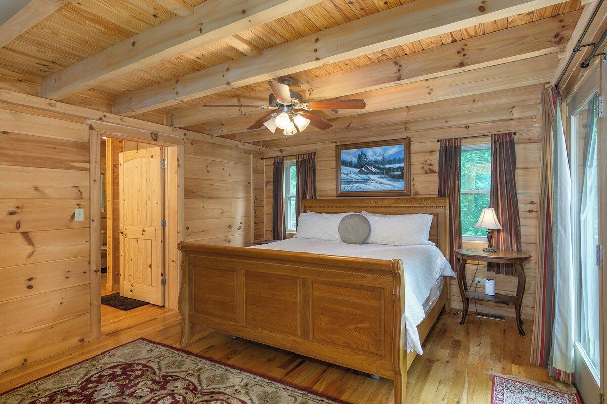 Ein Schlafzimmer einer Holzhütte mit einem Bett, einem Deckenventilator, einem Fenster mit Vorhängen und einem Bild an der Wand.
