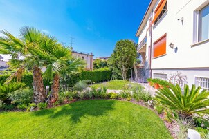 Un jardín cuidado con palmeras y diversas plantas, un césped impecable y un edificio moderno con fachada blanca y contraventanas naranjas.