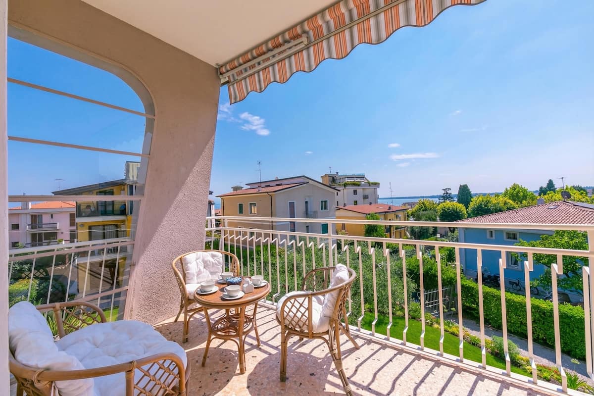 A balcony with wicker furniture, a table with cups and saucers, and a view of neighboring buildings and greenery.